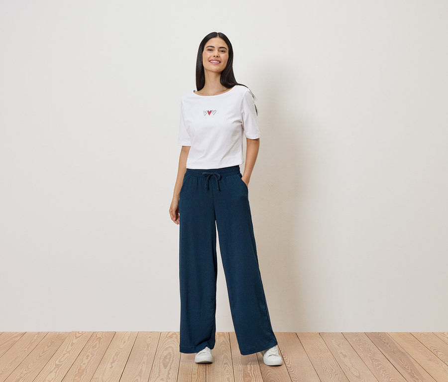 Frau mit weißem T-Shirt mit Herz-Motiv und weiter, dunkelblauer Hose mit Kordelzug. Sie trägt weiße Turnschuhe.