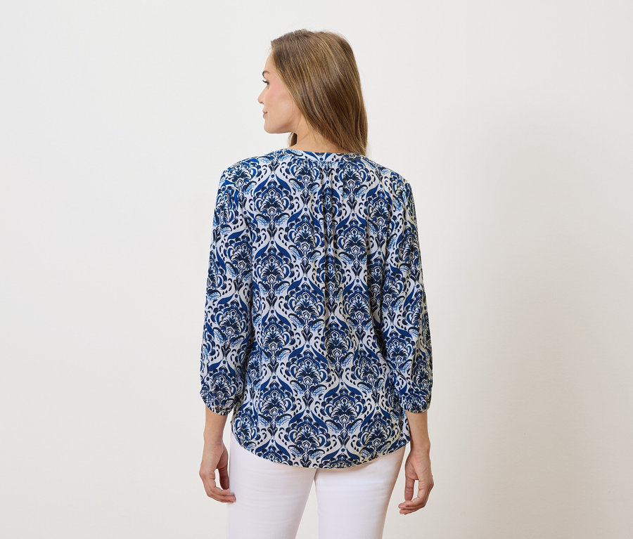 Eine Frau mit langen braunen Haaren trägt eine blau-weiß gemusterte Bluse und weiße Hosen.