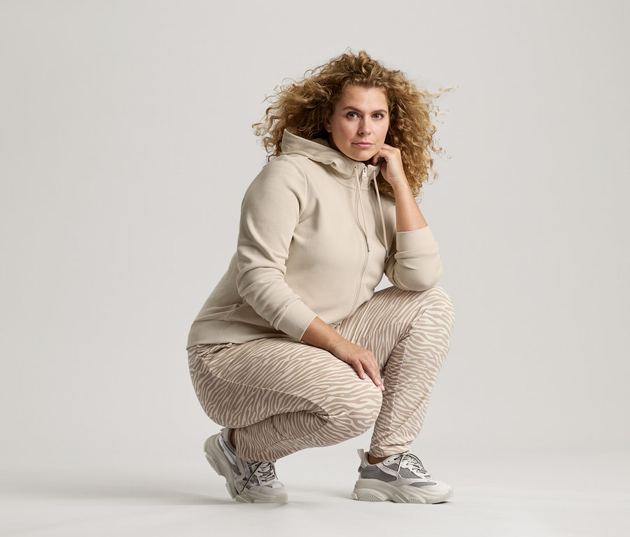 Frau mit lockigem Haar, die in beiger Jacke, Hose mit Zebramuster und Turnschuhen hockt und die Hand am Kinn hat.