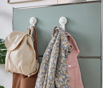 Eine beige Stofftasche, eine braune Wildledertasche, ein blaues geblümtes Hemd und eine rosa Jeansjacke hängen an weißen Saugnapf-Haken an einer mintgrünen Schrankwand.