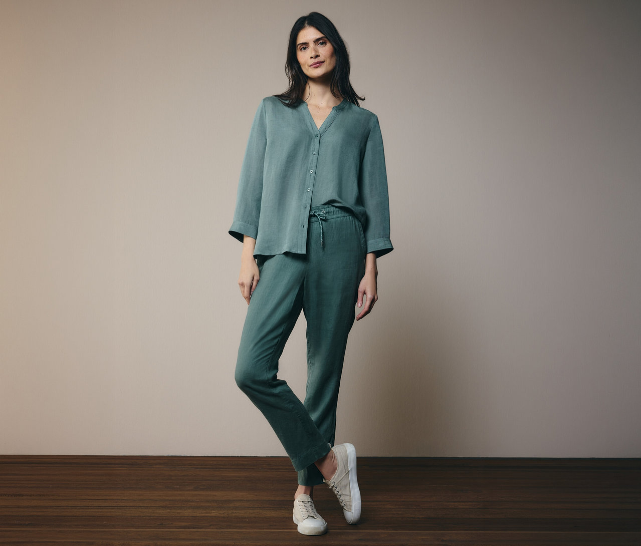 Frau in grüner Knopfbluse und Hose, ein Bein über das andere gekreuzt, dazu cremefarbene Schuhe.