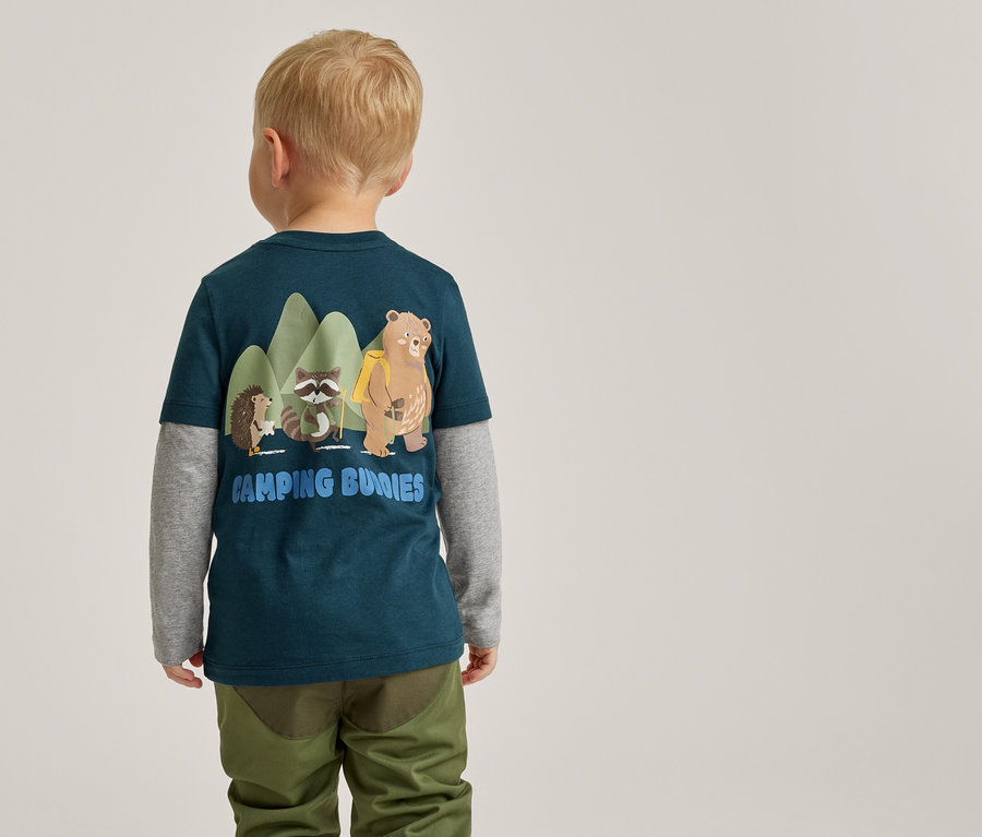 Ein kleines Kind, von hinten gesehen, trägt ein dunkelpetrolfarbenes T-Shirt über einem grauen Langarmshirt und olivgrüne Hosen. Das T-Shirt zeigt eine Grafik mit einem Igel, einem Waschbären und einem Bären vor grünen Bergen sowie den Text 'CAMPING BUNNIES'.