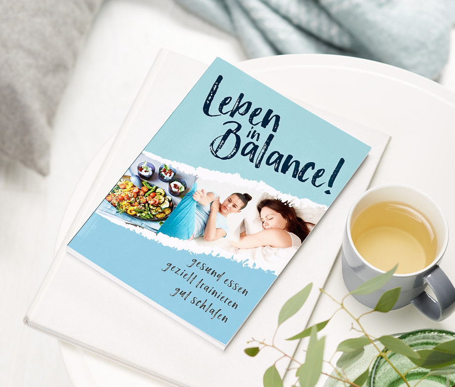 Auf einem weißen Tisch liegen das Buch "Leben in Balance", eine Tasse Tee und eine Vase mit Pflanzen.