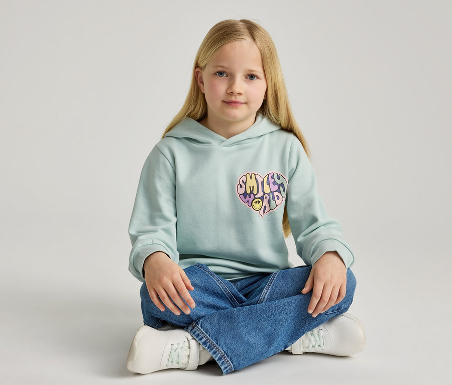 Mädchen sitzt mit gekreuzten Beinen und trägt einen hellblauen Hoodie mit einem Smiley-Herz-Aufdruck, Jeans und weiße Turnschuhe.