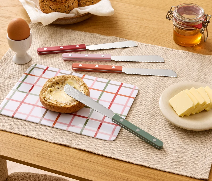 Auf einem Tisch liegen ein belegtes Brötchen, vier Messer, Butter, Honig, ein Ei und Brot.