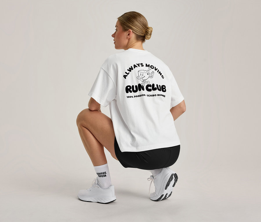 Eine Frau mit blondem Dutt hockt, von hinten gesehen. Sie trägt ein weißes, übergroßes Sportshirt mit schwarzem 'ALWAYS MOVING RUN CLUB'-Aufdruck am Rücken, schwarze Shorts, weiße Socken und weiße Sportschuhe.