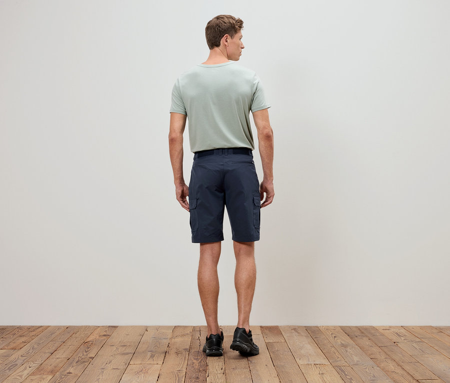 Mann von hinten in olivgrünem T-Shirt und blauen Cargoshorts, steht auf einem Holzboden vor einer weißen Wand.