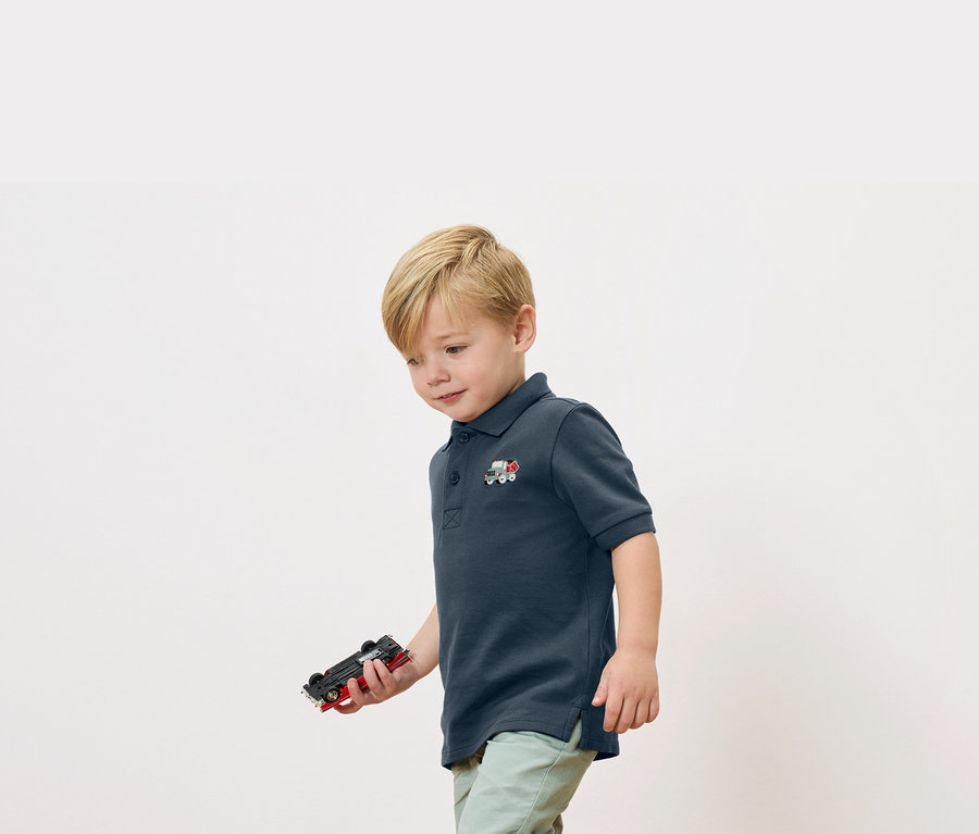 Ein blonder Junge hält ein Spielzeugauto in der Hand und trägt ein marineblaues Poloshirt mit einer kleinen Zugapplikation.