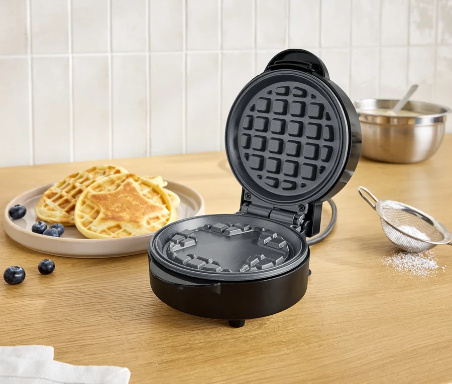 Offener, schwarzer Waffelautomat neben einem Teller mit Waffeln und Blaubeeren auf einem Holztisch.