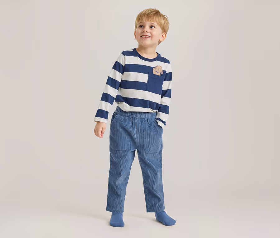 Junger Junge steht, blickt lächelnd nach oben und trägt ein blau-weiß gestreiftes Langarmshirt mit Tier-Brusttasche, blaue Cordhose und blaue Socken.
