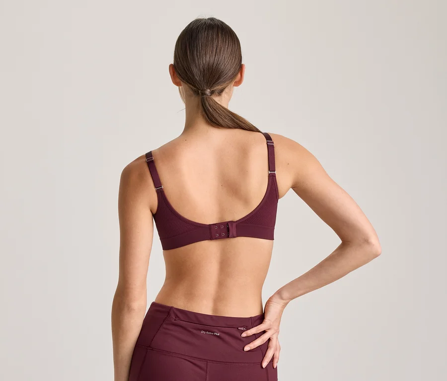 Frau von hinten, trägt einen bordeauxroten Sport-BH und passende Leggings. Ihr Haar ist zu einem Pferdeschwanz gebunden, ihre linke Hand liegt auf ihrer Hüfte.
