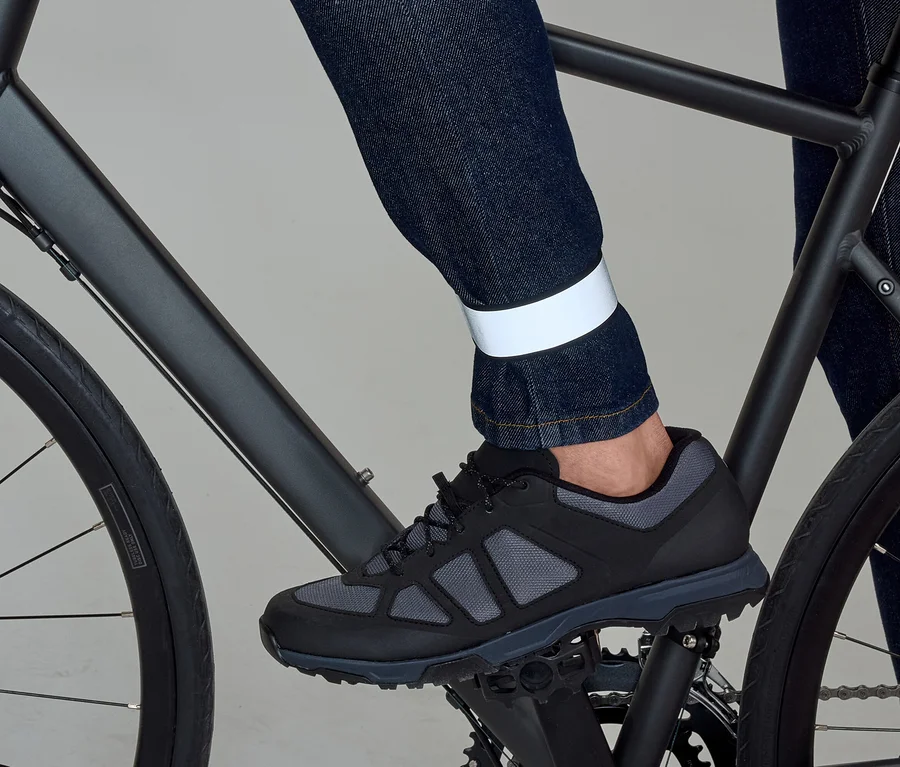 Ein Bein in dunkler Jeans mit reflektierendem Band am Knöchel tritt in einem schwarz-grauen Schuh ein schwarzes Fahrradpedal.
