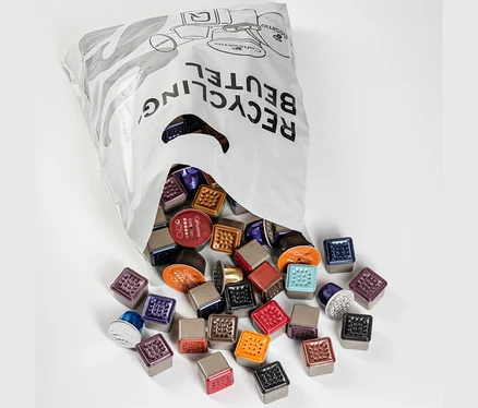 Ein offener Kostenfreier Recycling Sammelbeutel liegt auf weißem Untergrund, aus dem bunte Kaffeekapseln herausfallen.