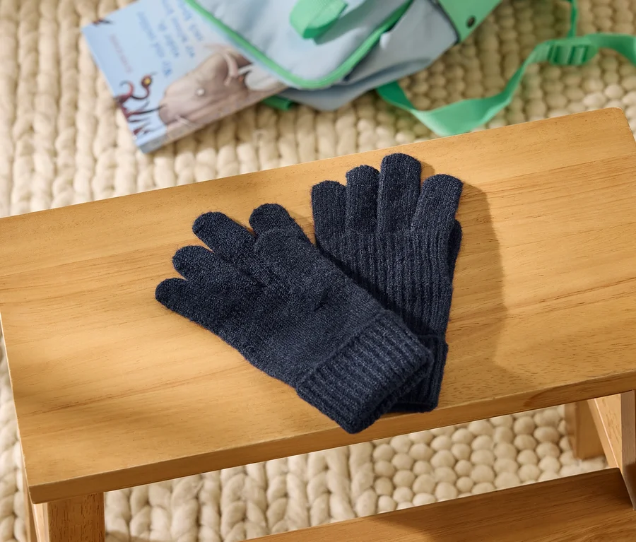 Dunkelblaue Strickhandschuhe liegen auf einem hellen Holzschemel. Im Hintergrund sind ein cremefarbener grob gestrickter Teppich, eine hellblaue Tasche und ein Buch mit Elefantenillustration zu sehen.