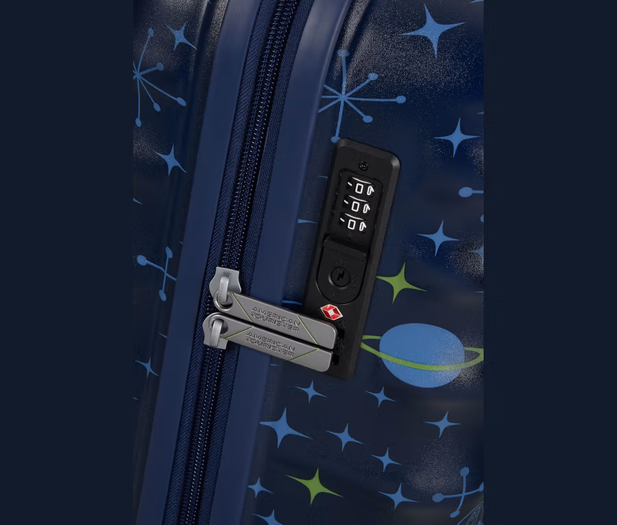 Nahaufnahme eines dunkelblauen Koffers mit Weltraum-Muster aus blauen und grünen Sternen und einem blauen Planeten. Der Koffer hat einen dunkelblauen Reißverschluss, zwei silbergraue Reißverschlusszipper mit „AMERICAN TOURISTER“-Branding und ein schwarzes Zahlenschloss mit TSA-Schlüsselloch.