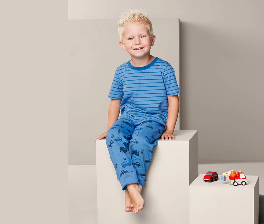 Ein Junge sitzt auf einem weißen Würfel und trägt einen blauen 3-teiligen Pyjama mit Autoaufdruck.