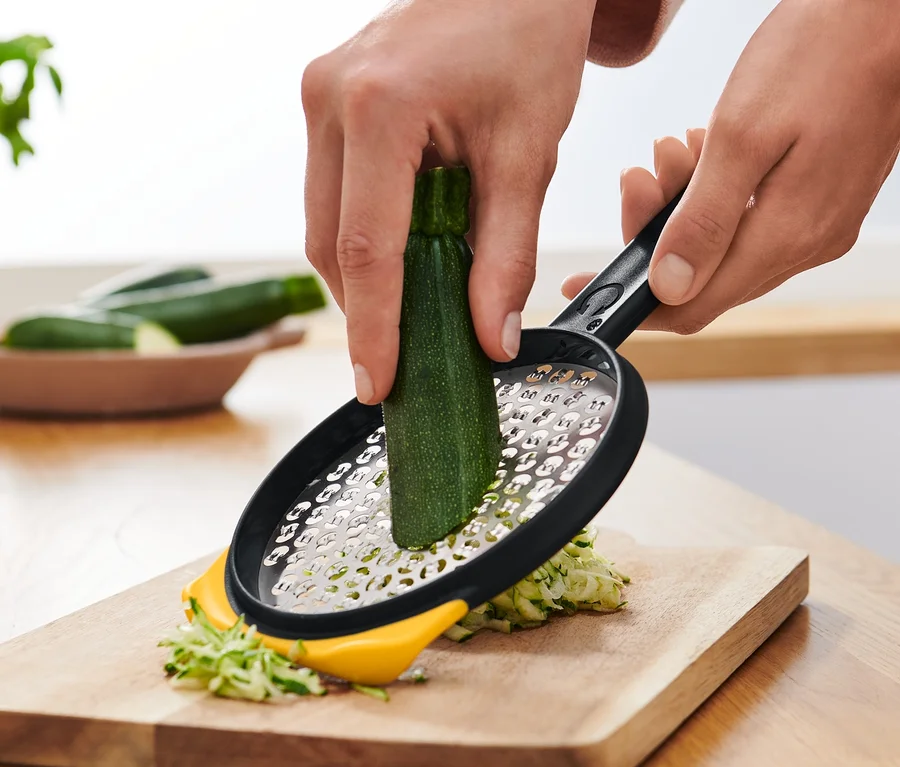Eine Person reibt Zucchini mit einer 2-seitigen Schnellreibe auf einem Holzbrett.