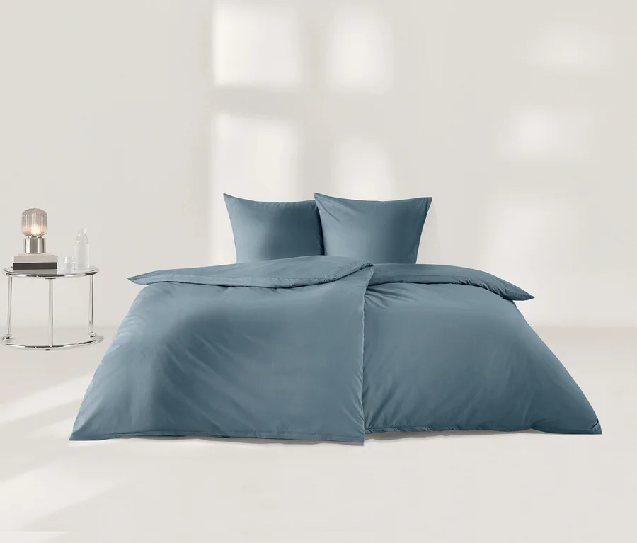 Blaue Perkal-Bettwäsche mit einem Bettbezug und zwei Kissen liegt auf einem Bett. Links steht ein runder Beistelltisch mit einer Lampe, einem Buch und einer Flasche.