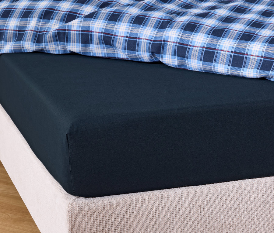 Detailaufnahme eines marineblauen Spannbettlakens auf einem Bett mit blau-weiß karierter Decke.
