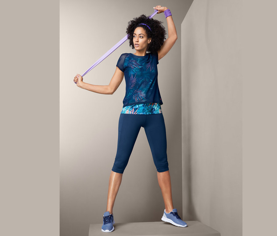 Frau hält ein lila Widerstandsband über ihren Kopf. Sie trägt ein Sportshirt mit Print, blaue Leggings und Turnschuhe.