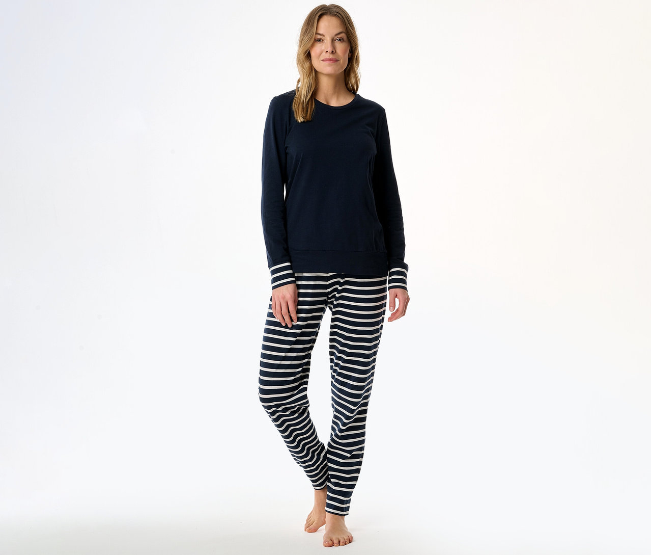 Frau in einem SCHIESSER Pyjama-Set, bestehend aus einem dunkelblauen Langarmshirt mit gestreiften Ärmelbündchen und einer dunkelblau-weiß gestreiften Hose, barfuß stehend.