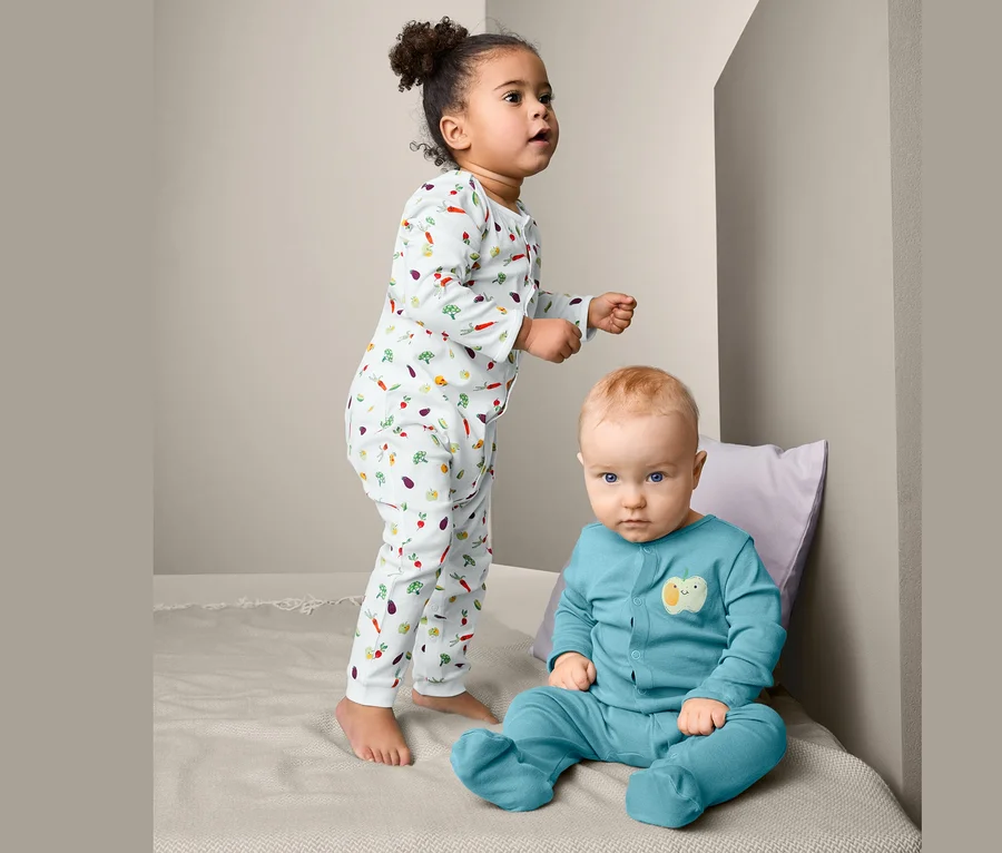 Zwei Kinder, ein Mädchen steht und ein Junge sitzt auf dem Bett. Das Mädchen trägt einen 2 Pyjamas mit Gemüseaufdruck, der Junge trägt einen blauen Pyjama.