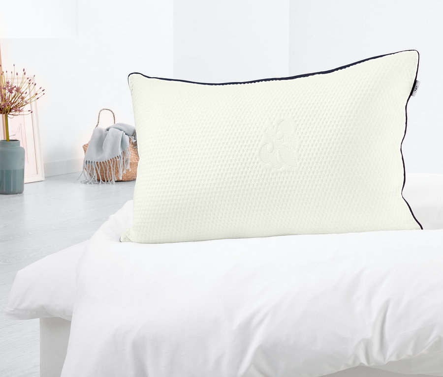 Ein weißes irisette®-Komfort-Schlafkissen mit dunkelblauer Paspel liegt auf einem weißen Bett.