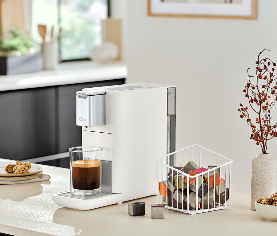 Eine Qbo ESSENTIAL Bright Sand Kaffeemaschine steht neben einem Glas Kaffee auf einer Küchentheke.