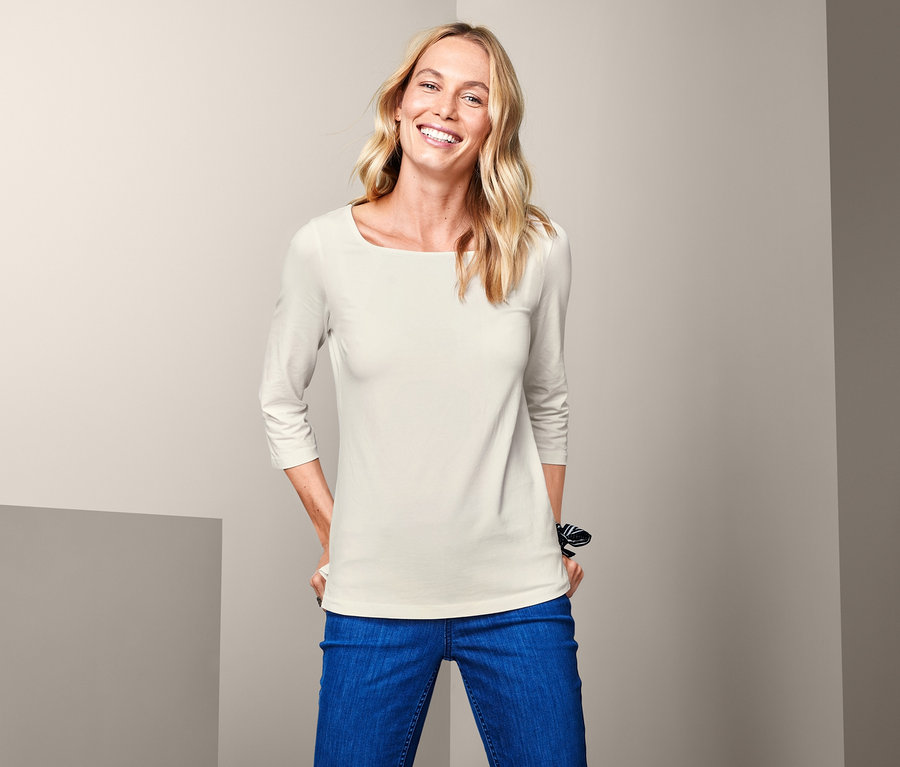 Frau mit blonden Haaren posiert in einem cremefarbenen Shirt mit 3/4-Arm und blauen Jeans.