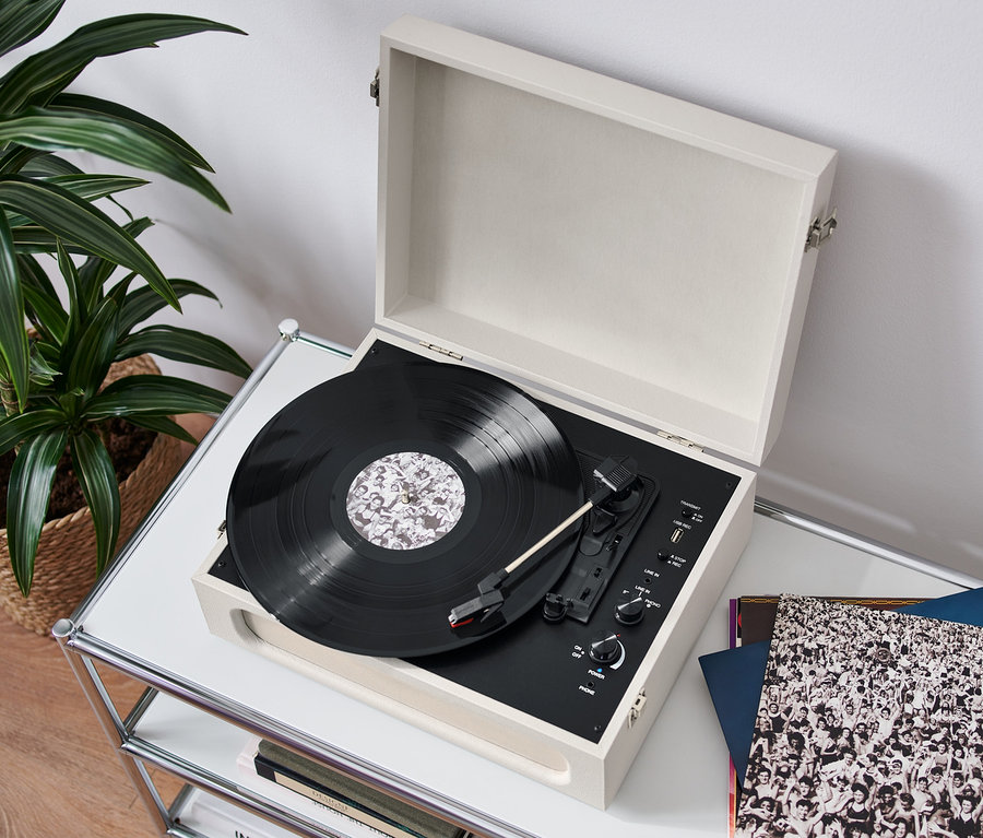 Ein geöffneter, tragbarer Retro-Plattenspieler mit Bluetooth®-Funktion steht auf einem weißen Tisch und spielt eine Schallplatte ab. Daneben liegen Schallplatten, im Hintergrund eine Pflanze im Korb.