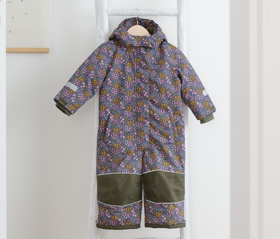 Ein Schneeanzug mit Blümchen-Print hängt auf einer weißen Leiter, daneben liegen gelbe Stiefel.