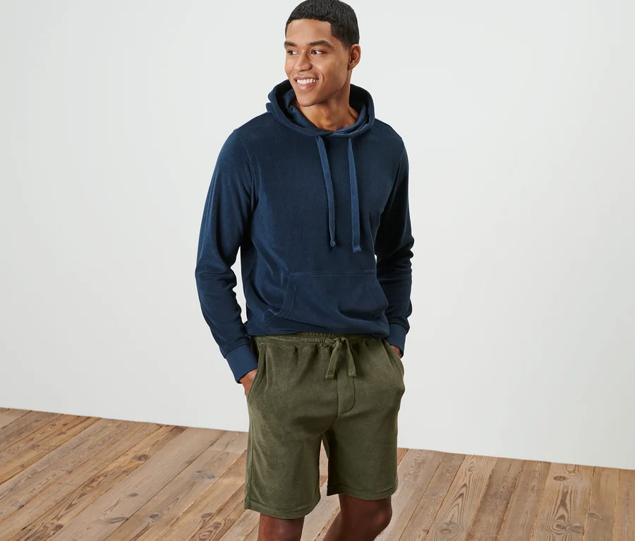 Ein lächelnder Mann steht und trägt einen blauen Kapuzenpullover und grüne Frottee-Shorts.