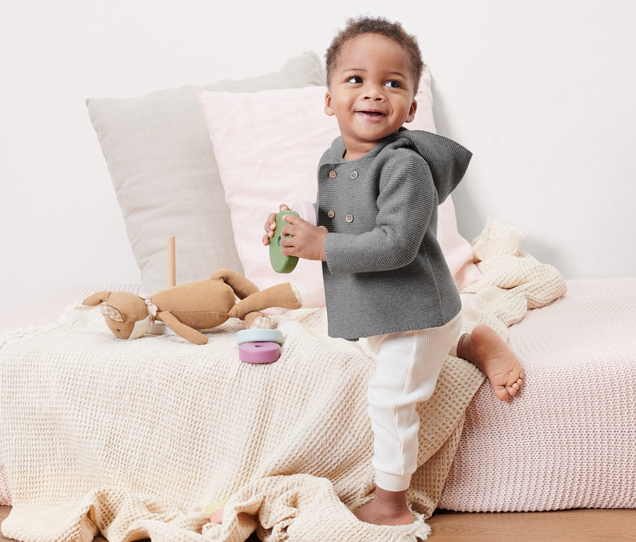 Ein Baby steht auf einem Bett und hält ein grünes Spielzeug. Es trägt eine grau melierte Baby-Strickjacke.
