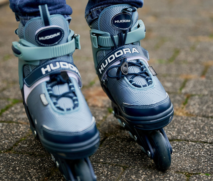 Nahaufnahme von den Füßen einer Person in blauen Jeans und schwarzen Knieschonern, die auf einem Bürgersteig stehen und graublaue HUDORA Leon 2.0 Inlineskates tragen.