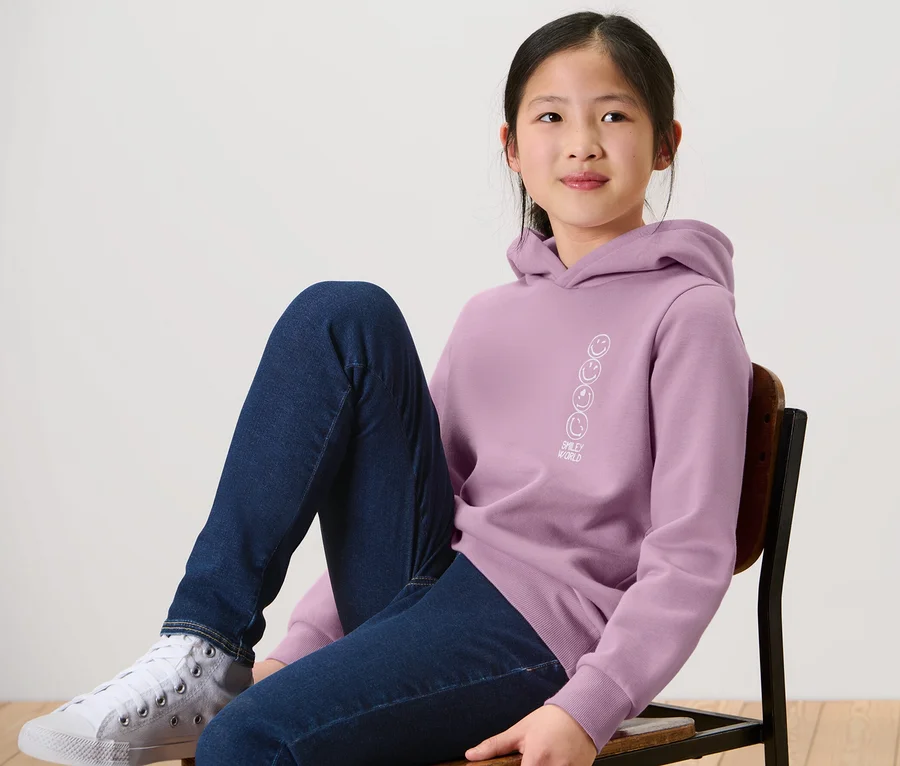 Mädchen sitzt auf einem Stuhl und trägt einen Kinder-Sweat-Hoodie »Smiley«.