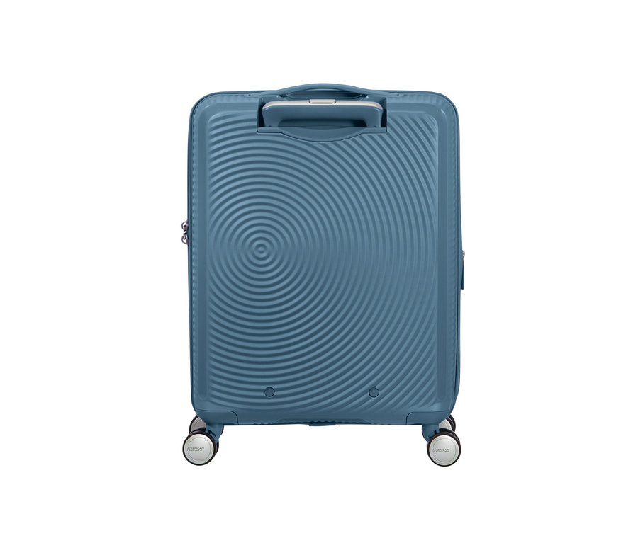 Ein kleiner, stone bluefarbener American Tourister Hartschalen-Koffer »Soundbox« Spinner steht auf Rollen.