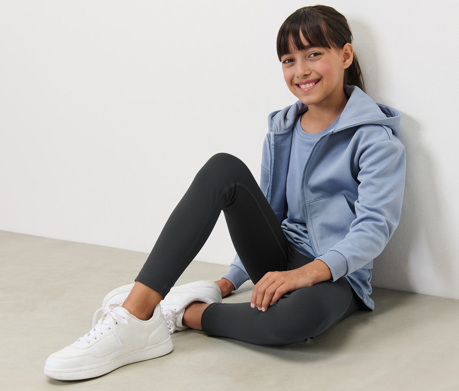 Lächelndes Mädchen sitzt auf dem Boden und trägt eine schwarze Kinder-Sporttight.