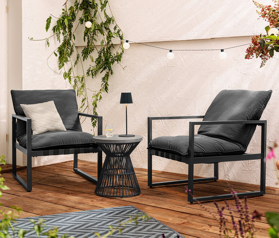 Zwei Relaxsessel stehen auf einer Terrasse neben einem Beistelltisch mit Polyzement-Tischplatte. Auf dem Tisch steht eine schwarze LED-Akku-Outdoor-Tischleuchte. Über den Sesseln hängt eine LED-Outdoor-Lichterkette.