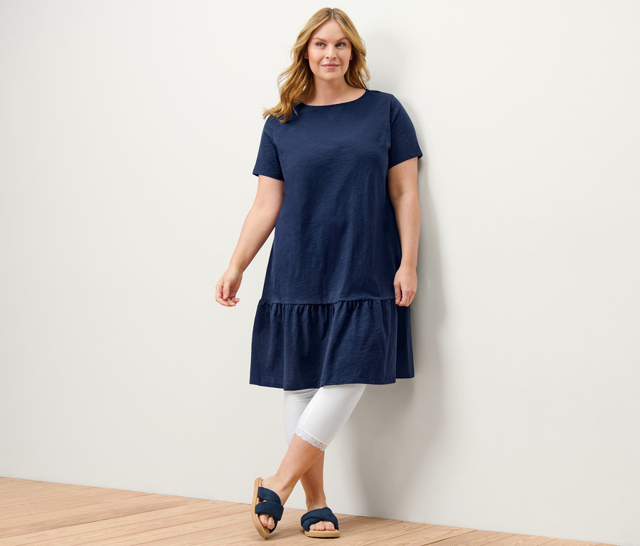 Eine Frau posiert in einem dunkelblauen Jerseykleid mit Volant und weißen 3/4-Leggings mit Spitzenborte.