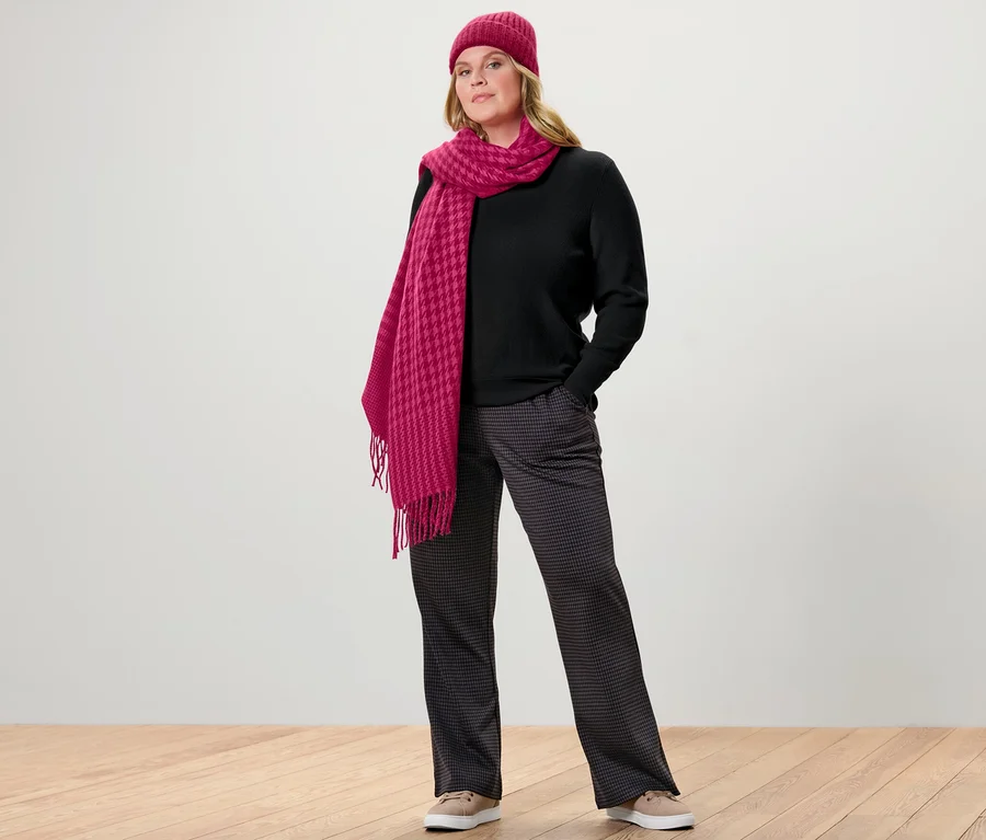 Frau posiert in Feinstrickpullover, schwarz, und Schlupfhose mit Hahnentritt-Jacquard. Sie trägt eine pinke Mütze und einen Schal.