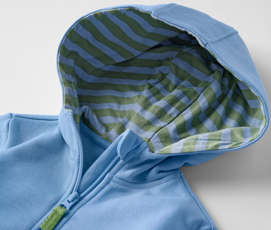 Detailansicht einer hellblauen Kinder-Sweatjacke mit Kapuze, deren Innenseite blau-grün gestreift ist, und einem Reißverschluss.