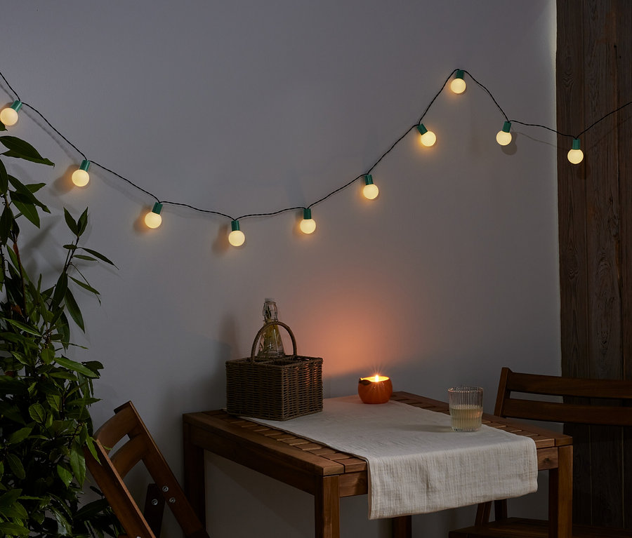 Eine LED-Lichterkette hängt an der Wand. Auf einem Tisch stehen ein Korb, eine Kerze und ein Glas.