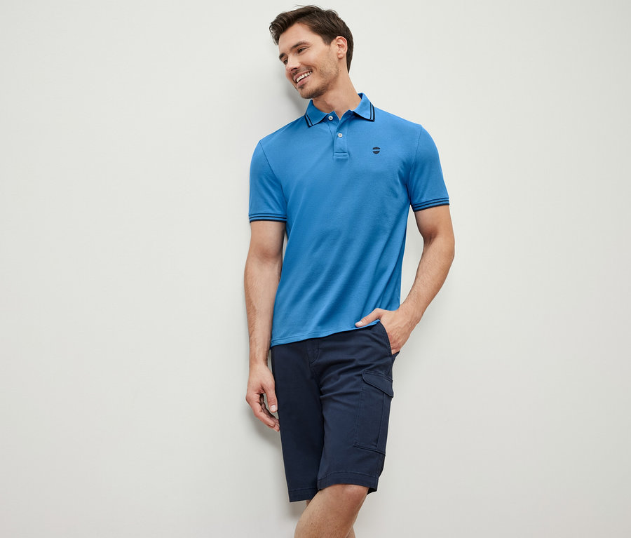 Mann an eine Wand gelehnt trägt ein blaues Piqué-Poloshirt und eine dunkelblaue Shorts.