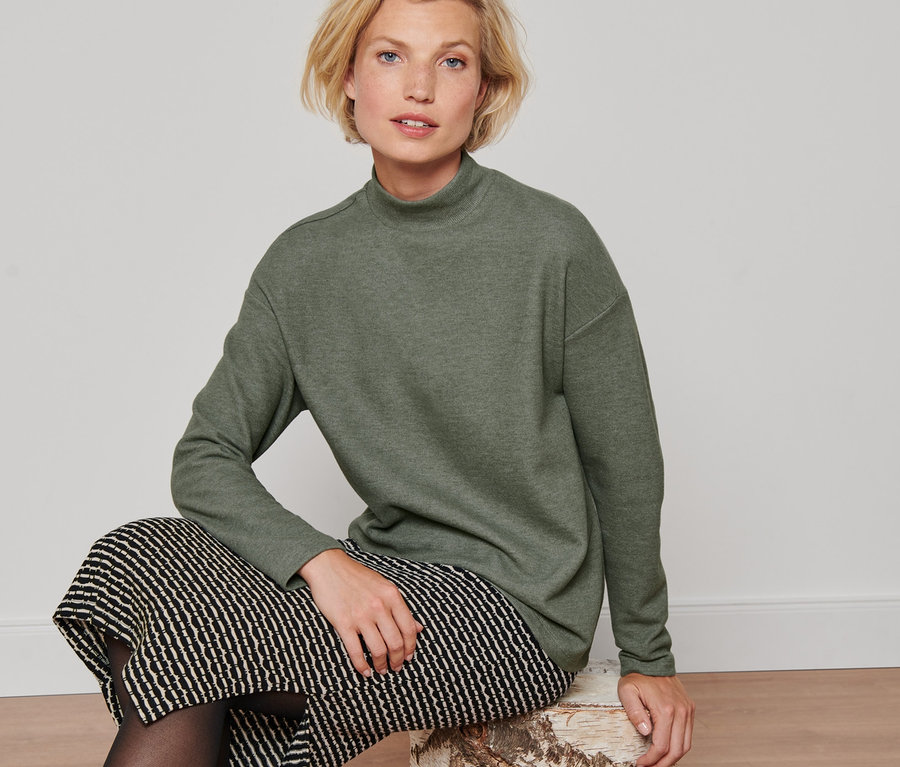 Frau sitzt auf einem Baumstumpf und trägt ein grünes Langarmshirt mit Stehkragen, einen schwarzen Rock und schwarze Stiefel.