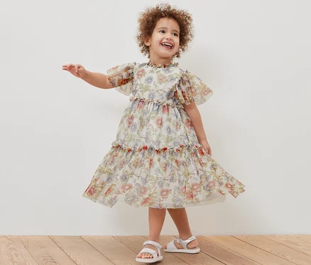 Ein lächelndes Mädchen posiert in einem geblümten Kinder-Tüllkleid und weißen Sandalen.