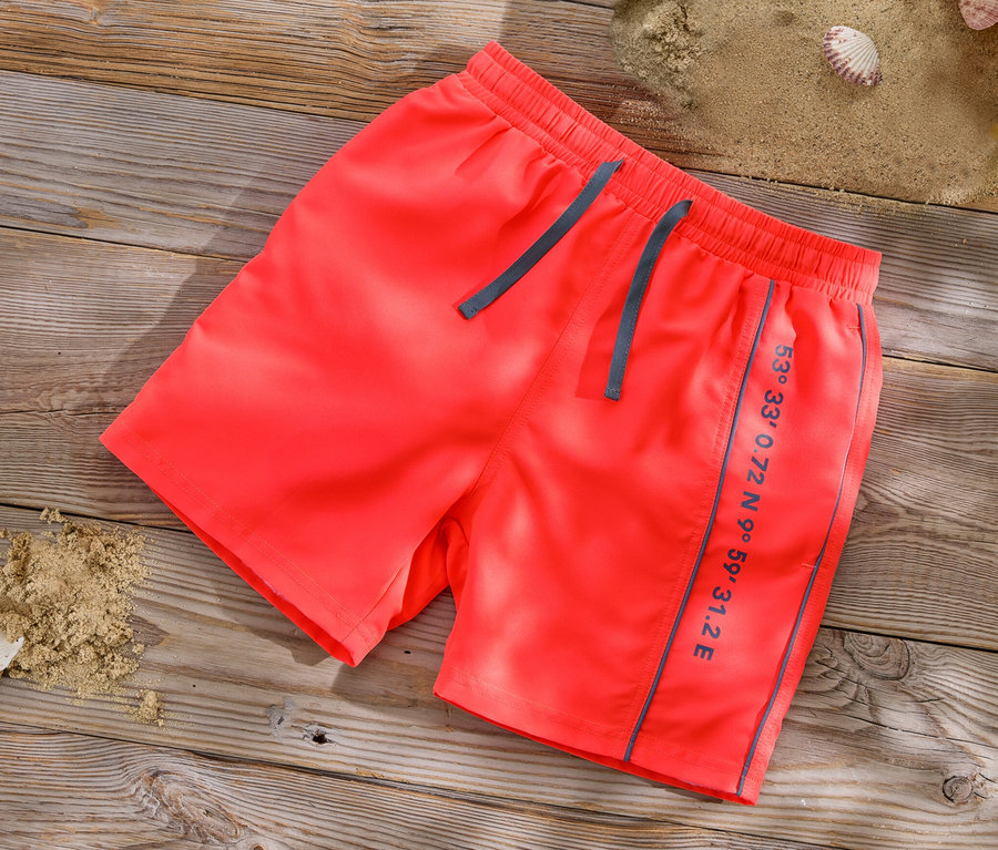Orangefarbene Kinder-Boardshorts liegen auf einer Holzoberfläche neben Sand, Muscheln und einem gelben Eimer.