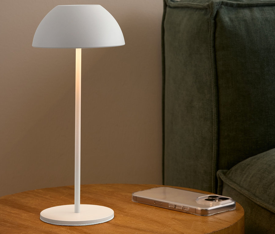 Eine weiße Lampe steht auf einem Holztisch neben einem Mobiltelefon und einem grünen Sofa.
