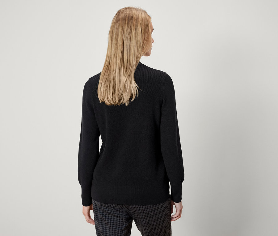 Von hinten ist eine Frau mit blonden Haaren zu sehen, die einen schwarzen Pullover und eine Hose mit Muster trägt.