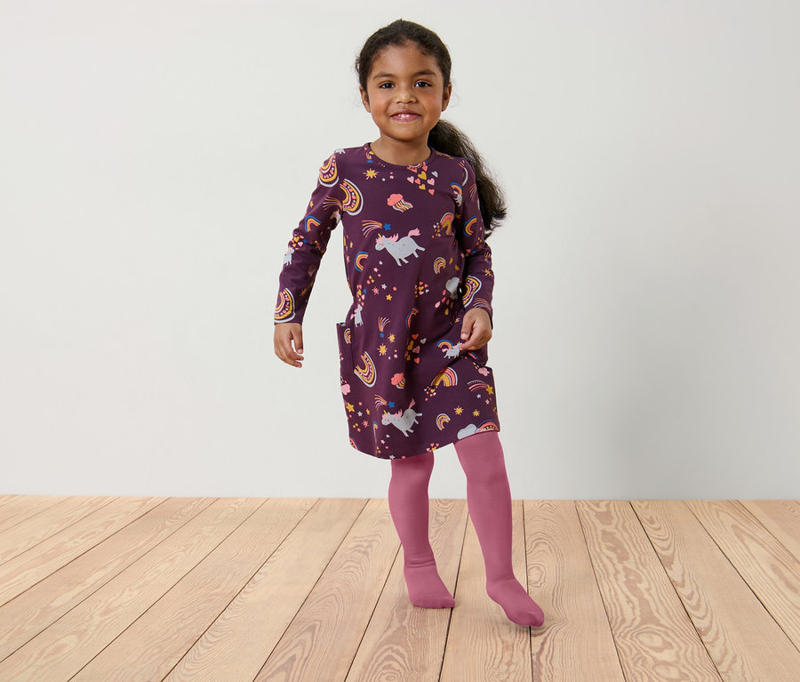 Mädchen in einem burgunderroten Kleid mit Einhorn- und Regenbogenmuster und rosa Strumpfhosen steht auf einem Holzboden.