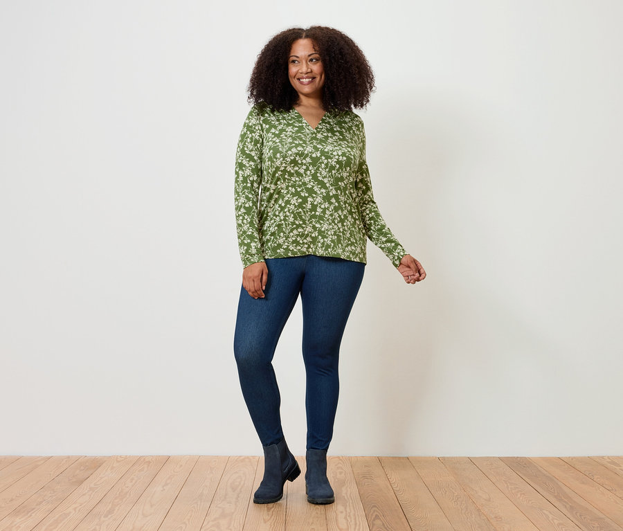 Eine Frau steht und posiert auf einem Holzboden, sie trägt ein grünes Oberteil mit Blumenmuster, Jeans-Leggings und Stiefel.